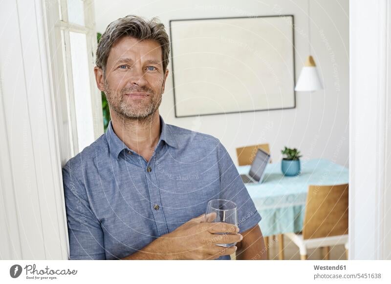 Mature man leaning against door case holding glass of water home ownership private owned home standing men males smiling smile Satisfaction Satisfied Content