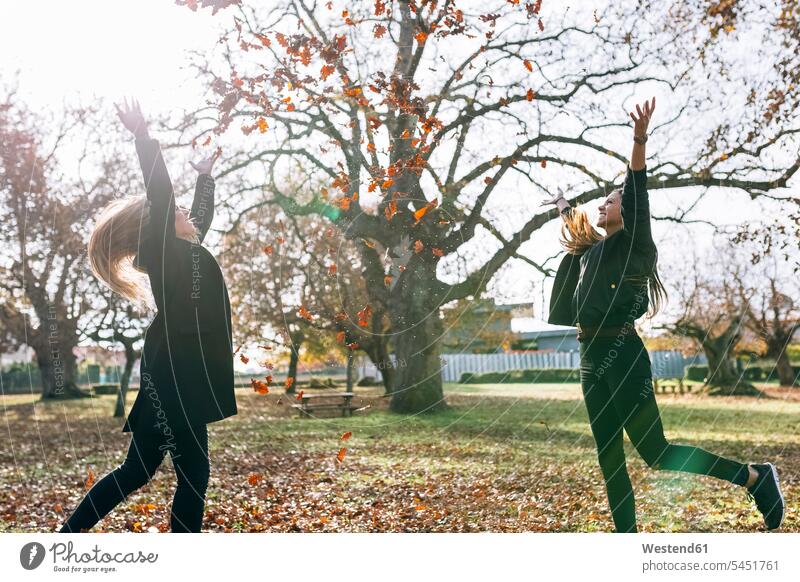 Two happy young women having fun in park in autumn parks woman females throwing Leaf Leaves female friends Adults grown-ups grownups adult people persons