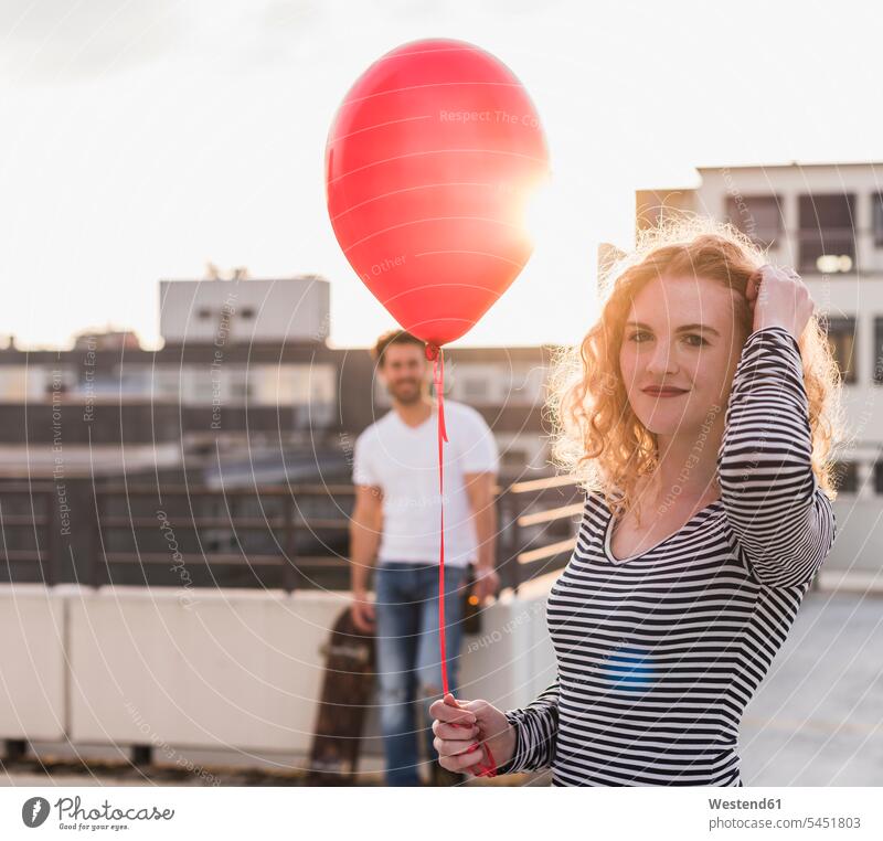 Portrait of smiling young wo man with red balloon on roof terrace at sunset balloons smile deck rooftop portrait portraits colour colours female skateboarder