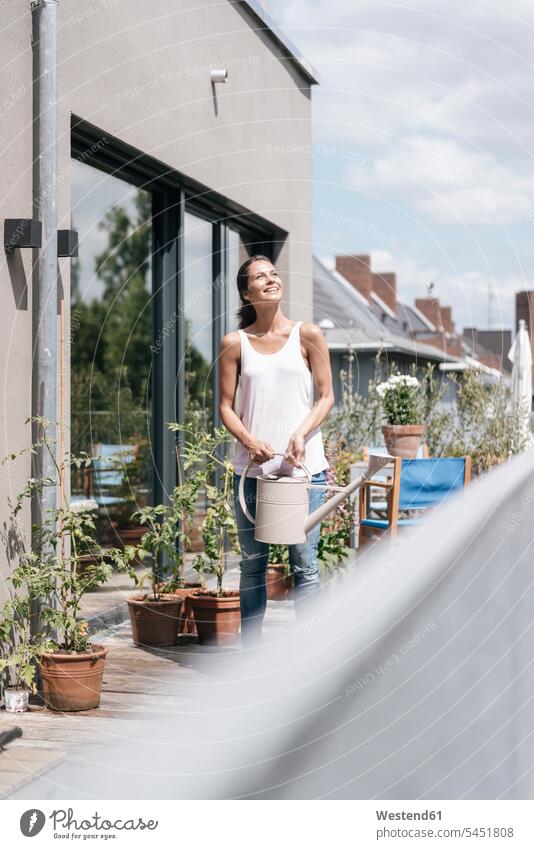 Smiling woman on balcony holding watering can watering cans balconies relaxed relaxation females women smiling smile relaxing Adults grown-ups grownups adult