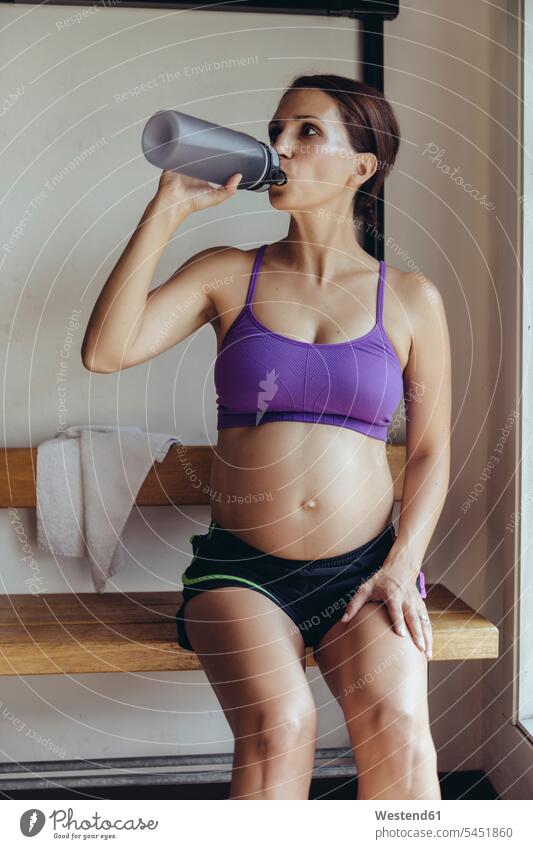 Sporty pregnant woman sitting in locker room drinking from bottle Locker Room Dressing Room females women Seated water bottle water bottles bottled water