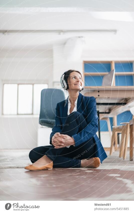 Smiling businesswoman sitting on the floor in a loft listening music with headphones headset females women businesswomen business woman business women Adults