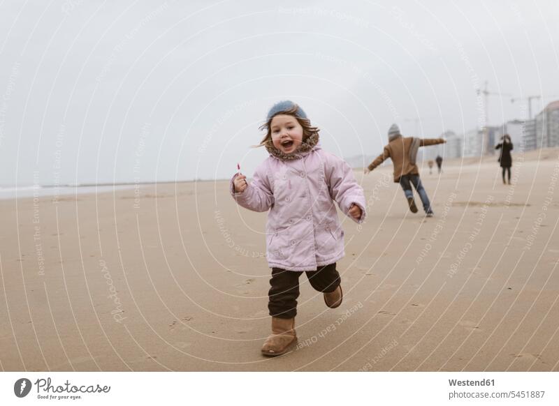 Happy little girl with lolly running on the beach in winter while her brother playing in the background happiness happy hibernal brothers Lollipop Lollipops