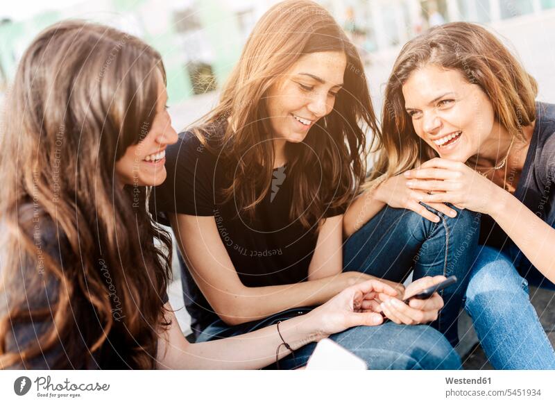 Three happy young women sitting outdoors looking at cell phone smiling smile female friends mobile phone mobiles mobile phones Cellphone cell phones mate