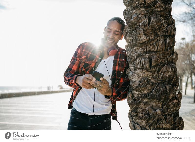 Spain, smiling young man with headphones leaning against palm tree trunk looking at cell phone Smartphone iPhone Smartphones headset men males mobile phone