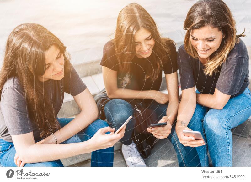 Three smiling young women sitting outdoors looking at cell phones female friends smile mobile phone mobiles mobile phones Cellphone mate friendship telephones