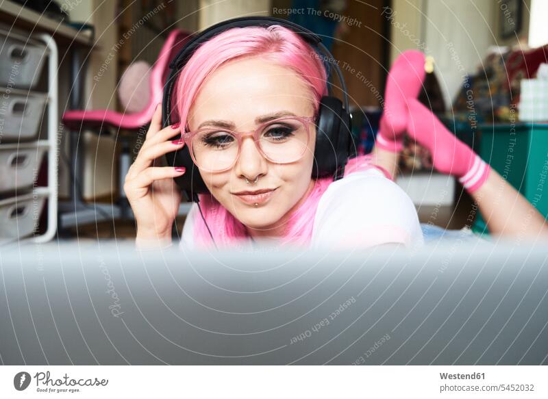 Young woman with pink hair listening to music via laptop at home females women smiling smile headphones headset glasses specs Eye Glasses spectacles Eyeglasses