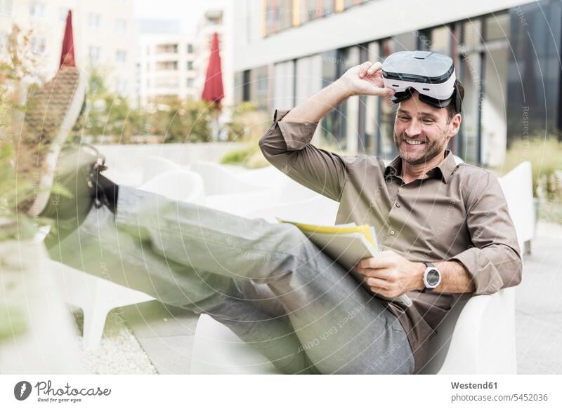 Smiling man with Virtual Reality Glasses and documents sitting on terrace Businessman Business man Businessmen Business men smiling smile freelancer freelancing