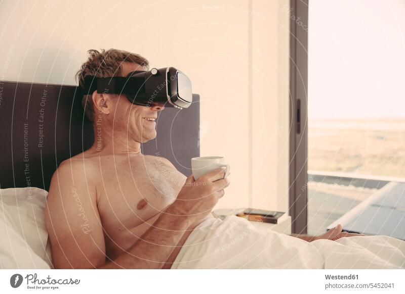 Man wearing VR glasses in bed holding cup of coffee smiling smile specs Eye Glasses spectacles Eyeglasses man men males Coffee apartment flats apartments beds