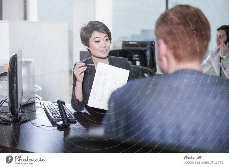 Woman showing sheet of paper to man at desk in office talking speaking offices office room office rooms workplace work place place of work business