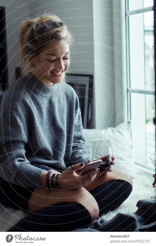 Smiling young woman with cup of coffee relaxing on sheepskin at home looking at cell phone females women Smartphone iPhone Smartphones Adults grown-ups grownups