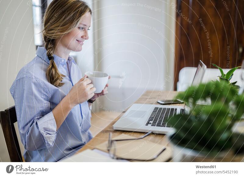 Smiling woman sitting at desk with cup of coffee and laptop desks Seated smiling smile Coffee Laptop Computers laptops notebook females women Table Tables Drink