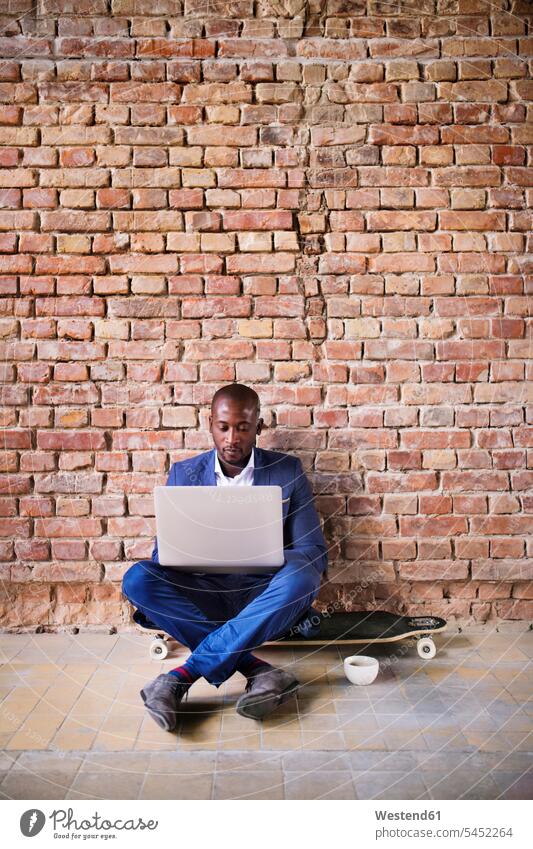 Businessman sitting on longboard at brick wall using laptop Laptop Computers laptops notebook Business man Businessmen Business men skateboard Skate Board