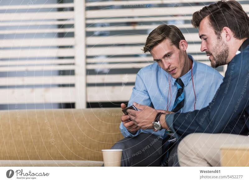 Two colleagues sharing cell phone in office lounge talking speaking sitting Seated offices office room office rooms mobile phone mobiles mobile phones Cellphone