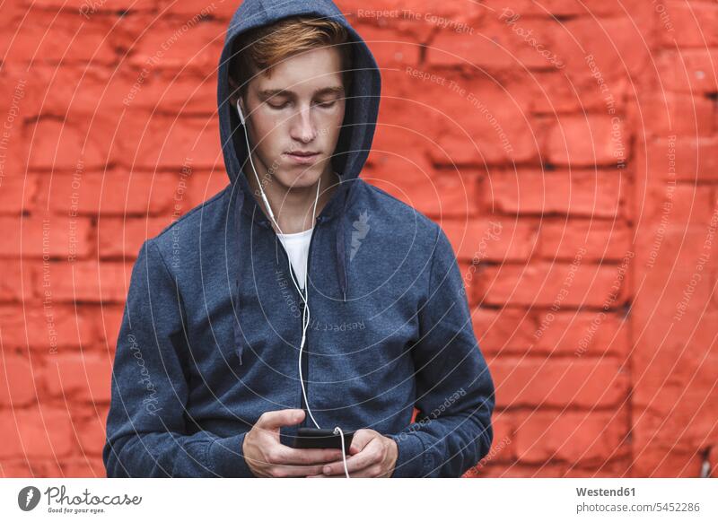 Young man with cell phone and earphones at red brick wall hearing men males ear phone ear phones mobile phone mobiles mobile phones Cellphone cell phones