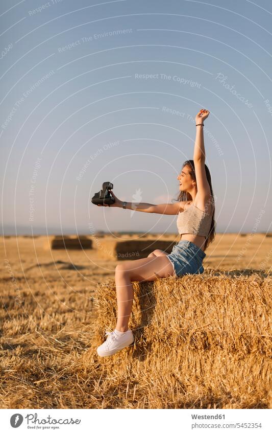 Young woman sitting on straw bale taking selfie with instant camera Selfie Selfies females women Adults grown-ups grownups adult people persons human being