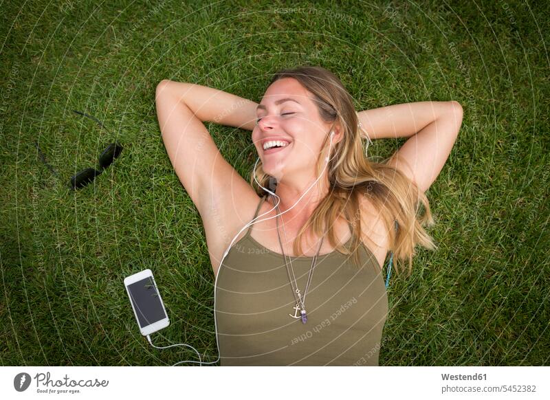 Happy young woman with earphones relaxing on lawn ear phone ear phones portrait portraits smiling smile relaxed relaxation park parks lying laying down lie