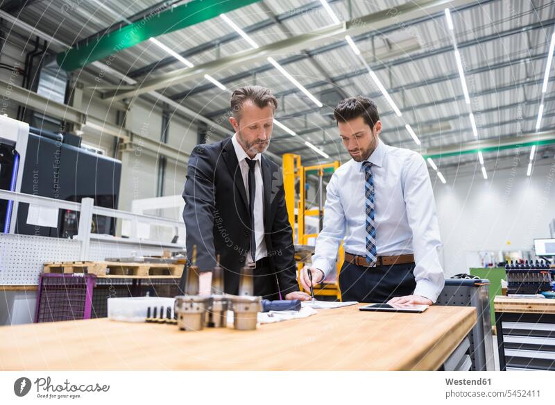 Two businessmen at table in factory shop floor examining product factories colleagues Businessman Business man Businessmen Business men tablet digitizer