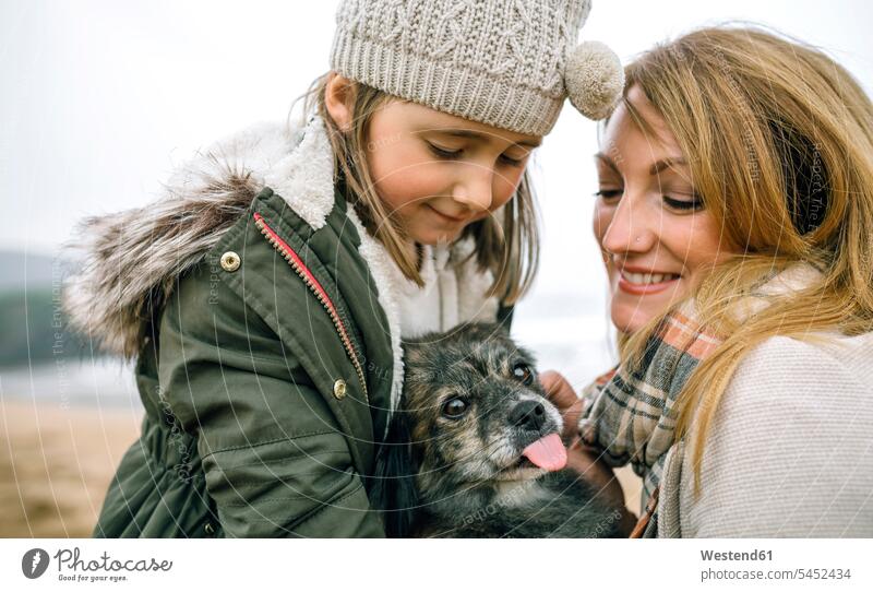 Mother and daughter stroking their dog on the beach in winter beaches daughters smiling smile dogs Canine child children family families people persons