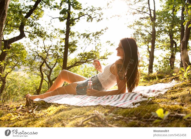 Smiling young woman in forest lying on blanket with drink and apple woods forests smiling smile relaxed relaxation females women relaxing Adults grown-ups