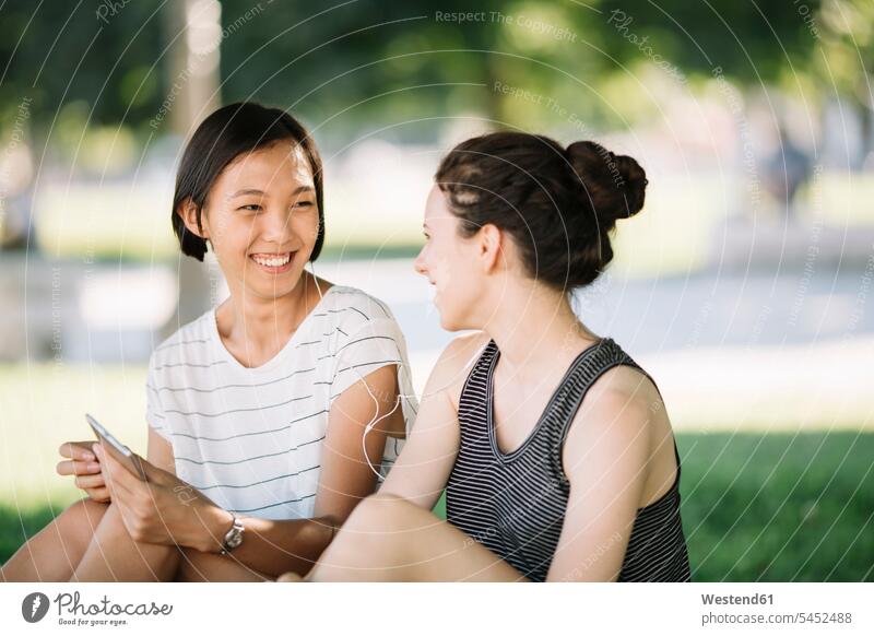Two friends sitting in city park listening music together with earphones female friends mate friendship multicultural City Park municipal park urban park