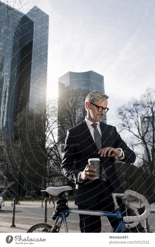 Grey-haired businessman with bicycle and coffee to go in the city looking at smartwatch Coffee bikes bicycles town cities towns Businessman Business man