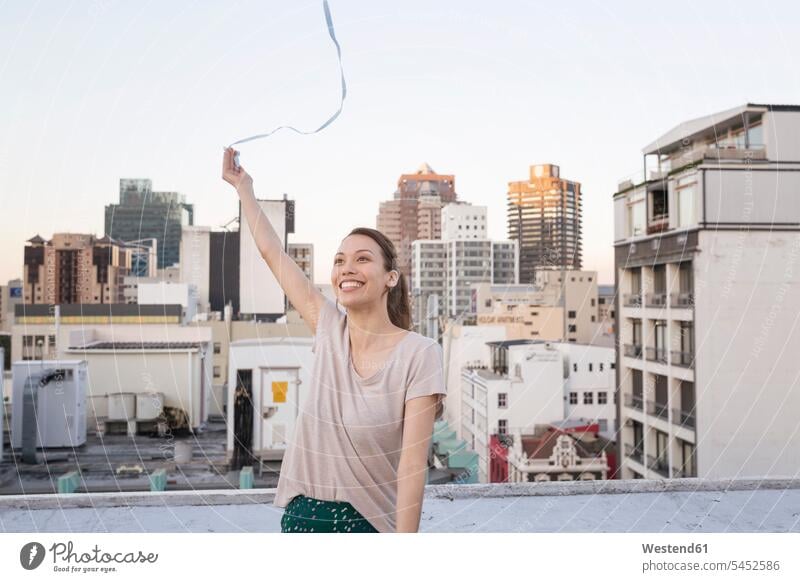 Young woman standing on a rooftop terrace, playing with a ribbon carefree roof terrace deck parapet balustrade females women happiness happy flying smiling