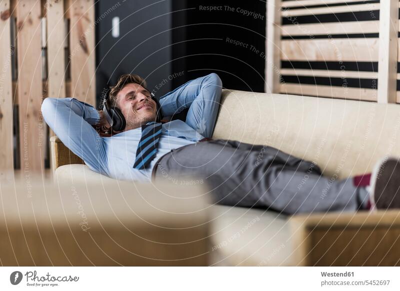 Businessman with headphones lying on couch in office lounge laying down lie lying down Business man Businessmen Business men headset relaxed relaxation