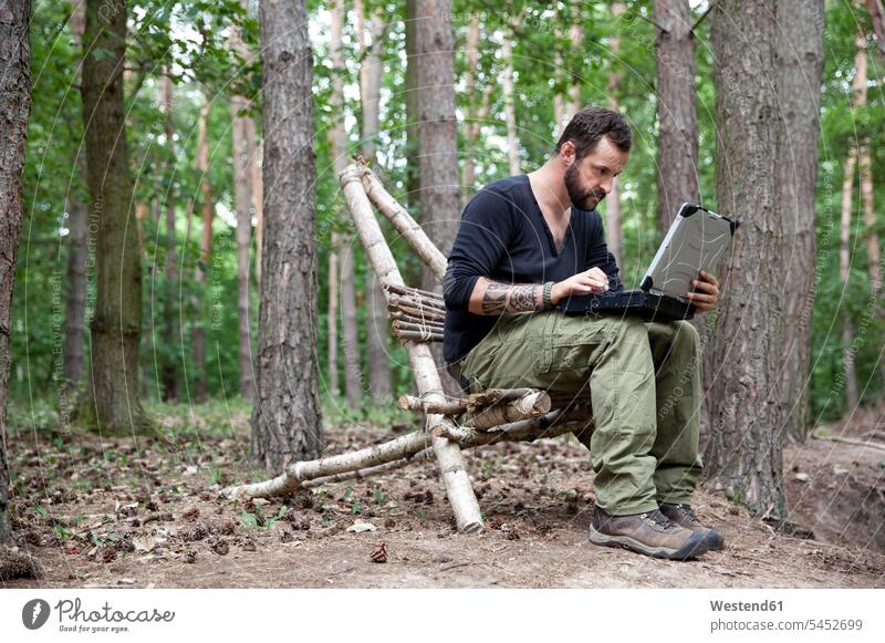 Man sitting on self-made wooden chair in forest using laptop Laptop Computers laptops notebook man men males woods forests Seated computer computers Adults