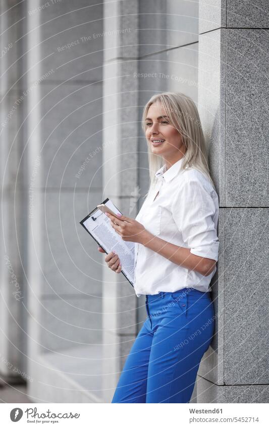Smiling businesswoman with clipboard and cell phone leaning against a wall smiling smile businesswomen business woman business women mobile phone mobiles
