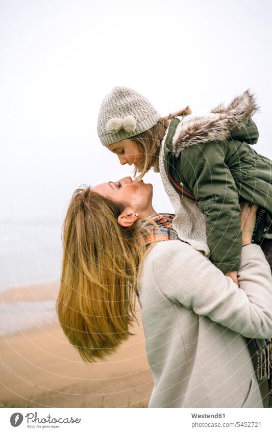 Woman lifting up and kissing daughter on the beach in winter daughters mother mommy mothers mummy mama beaches child children family families people persons