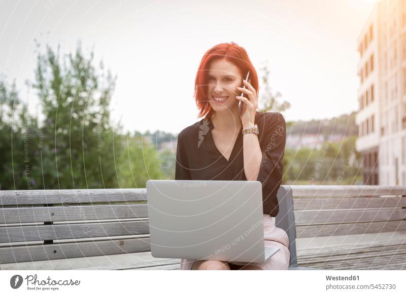 Young woman sitting on bench with her laptop, making a phone call Seated Smartphone iPhone Smartphones females women young on the phone telephoning