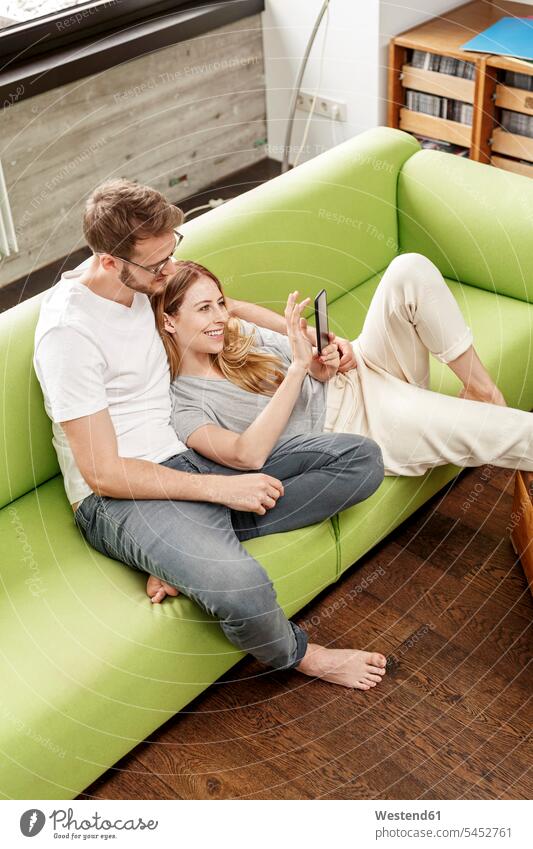 Smiling young couple on couch in living room at home sharing cell phone twosomes partnership couples mobile phone mobiles mobile phones Cellphone cell phones