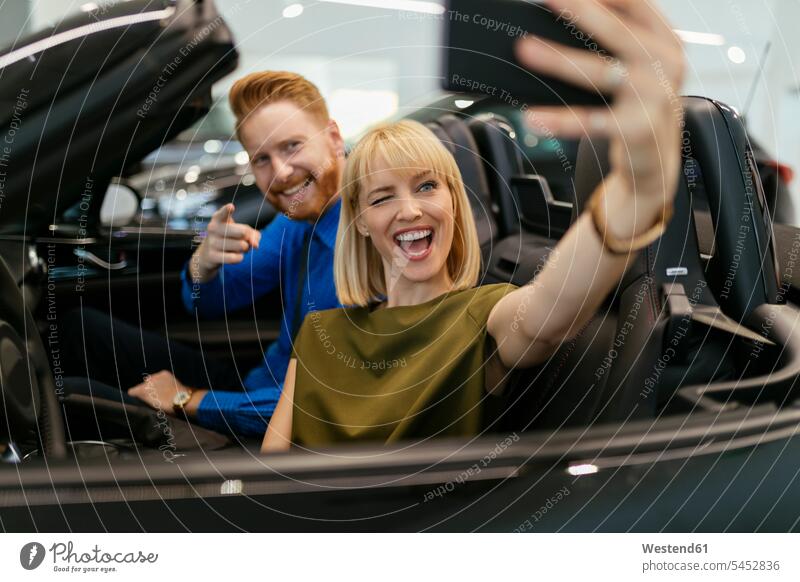 Couple taking selfies, sitting in convertible in car dealership automobile Auto cars motorcars Automobiles Selfie Selfies couple twosomes partnership couples