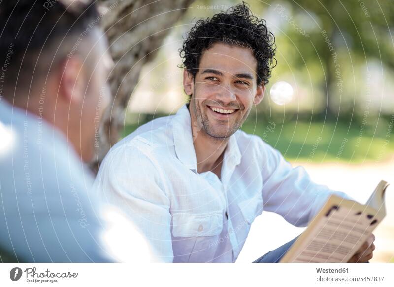 Smiling young man holding book looking at friend in park smiling smile books men males Adults grown-ups grownups adult people persons human being humans