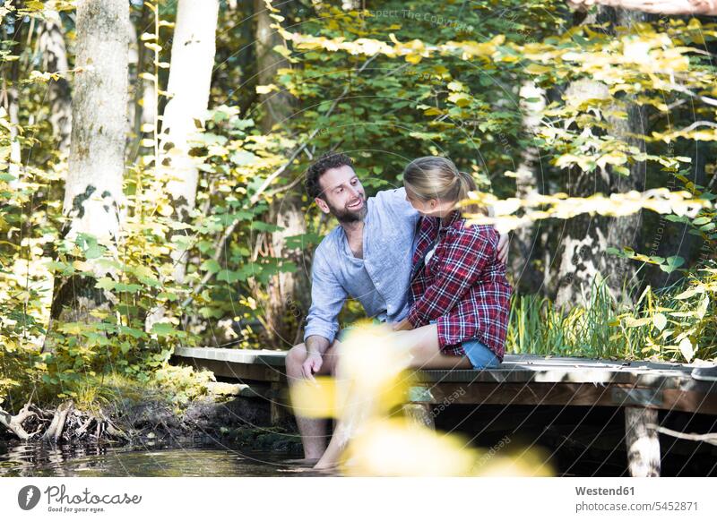 Young couple sitting on a bridge in forest with feet in water smiling smile twosomes partnership couples bridges brook brooks rivulet woods forests Seated