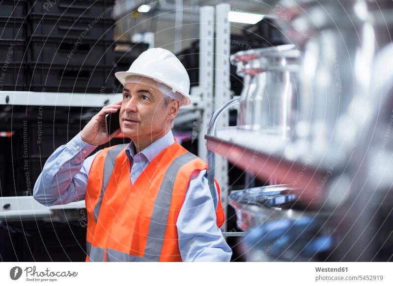 Man in factory hall wearing safety vest and hard hat talking on cell phone man men males on the phone call telephoning On The Telephone calling mobile phone