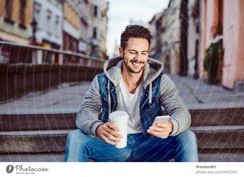 Man with coffee sitting on stairs in the city using phone man men males mobile phone mobiles mobile phones Cellphone cell phone cell phones Seated smiling smile