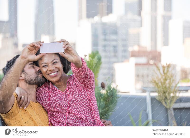 Couple sitting on rooftop terrace, taking smart phone selfies photographing smiling smile Seated couple twosomes partnership couples roof terrace deck people