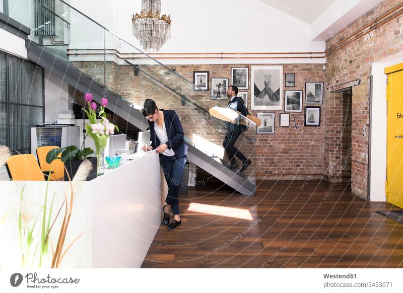 Businesswoman standing at reception while man with longboard is ascending stairs in background caucasian caucasian ethnicity caucasian appearance european