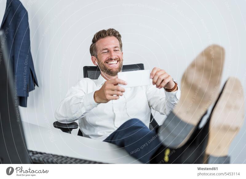 Businessman with earphones sitting at desk with feet up looking at cell phone Business man Businessmen Business men portrait portraits business people