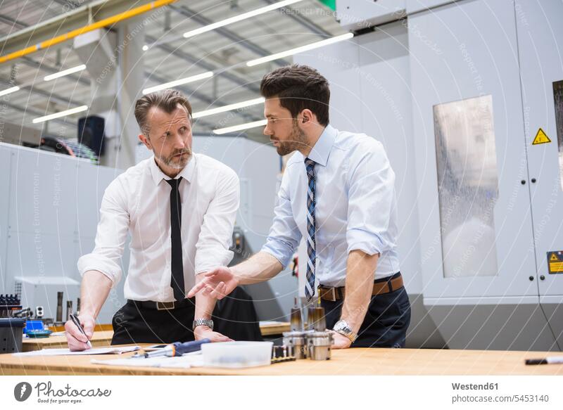 Two businessmen at table in factory shop floor discussing factories Businessman Business man Businessmen Business men colleagues business people businesspeople