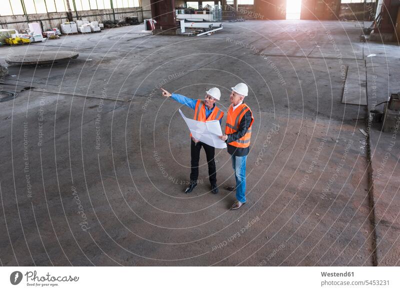Two men with plan wearing safety vests talking in old industrial hall construction plan building plan architectural drawing colleagues shop floor factory hall