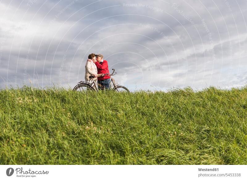 Senior couple with bicycles kissing in rural landscape under cloudy sky twosomes partnership couples senior men senior man elder man elder men senior citizen