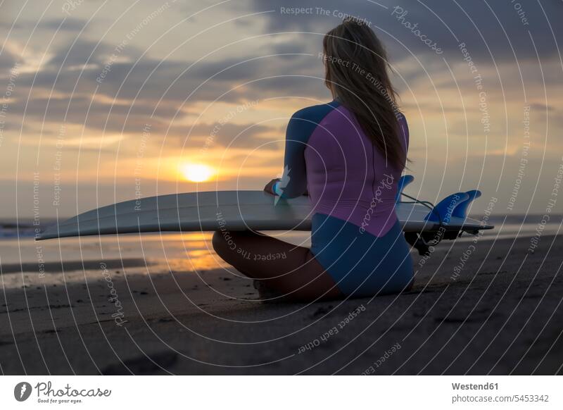 Indonesia, Bali, young woman with surfboard sitting on beach at sunset evening mood leisure free time leisure time surfboards females women Seated surfer
