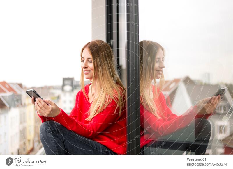 Smiling young woman sitting at the window in city apartment looking at cell phone smiling smile mobile phone mobiles mobile phones Cellphone cell phones Seated