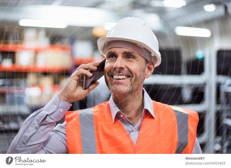 Smiling man in factory hall wearing safety vest and hard hat talking on cell phone men males on the phone call telephoning On The Telephone calling mobile phone
