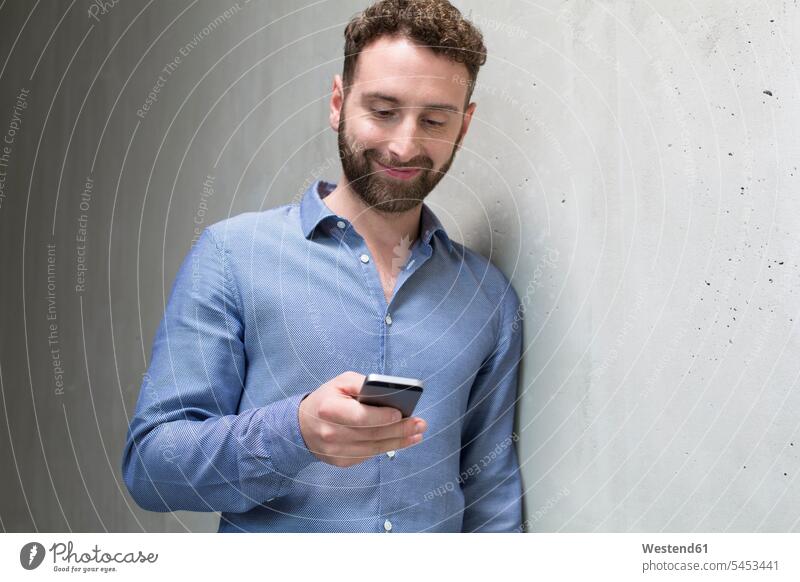 Smiling man looking at cell phone at concrete wall men males concrete walls mobile phone mobiles mobile phones Cellphone cell phones smiling smile Adults