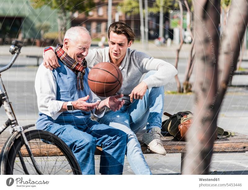 Senior man and adult grandson with basketball talking on a bench sitting Seated grandsons benches grandfather grandpas granddads grandfathers happiness happy
