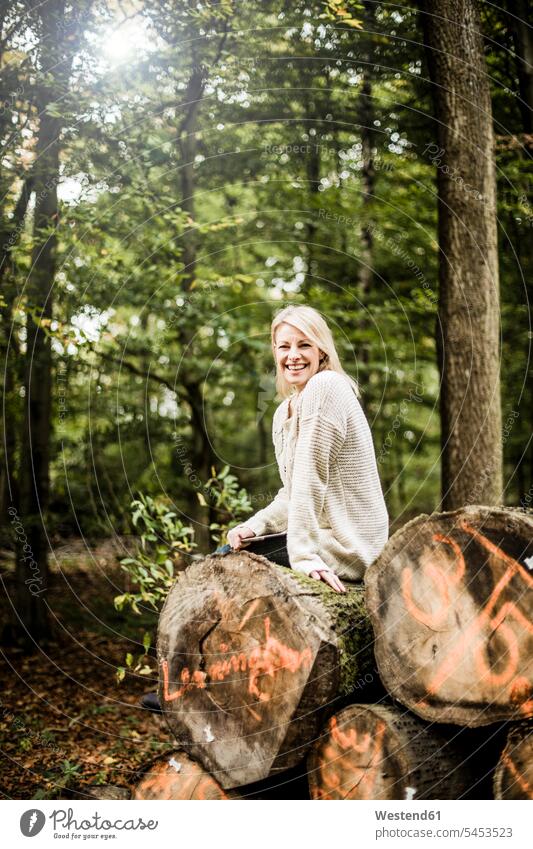 Portrait of happy woman sitting on stack of logs in the forest with tablet woods forests laughing Laughter females women portrait portraits positive Emotion